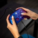 Mano femenina sujetando un mando de Xbox azul y morado con cable negro, botones coloridos y diseño ergonómico para juegos